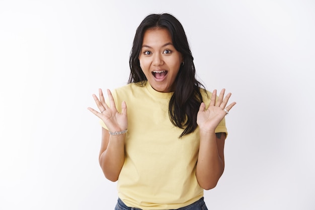Mädchen, das Überraschung macht, kommt heimlich zum Haus des Freundes, schreit Hallo und winkt mit erhobenen Handflächen, die fröhlich und fröhlich mit enthusiastischen Ausdrücken vor weißem Hintergrund in gelbem T-Shirt posieren