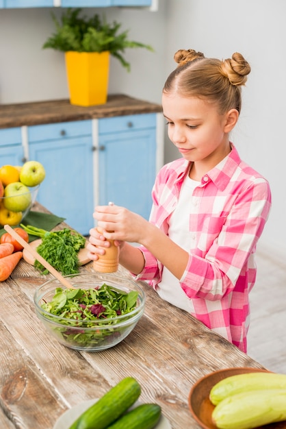 Mädchen, das den Pfeffer in der frischen Salatschüssel auf Holztisch hinzufügt