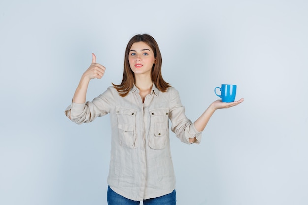 Mädchen, das eine tasse tee hält, daumen im hemd zeigt und zufrieden aussieht.