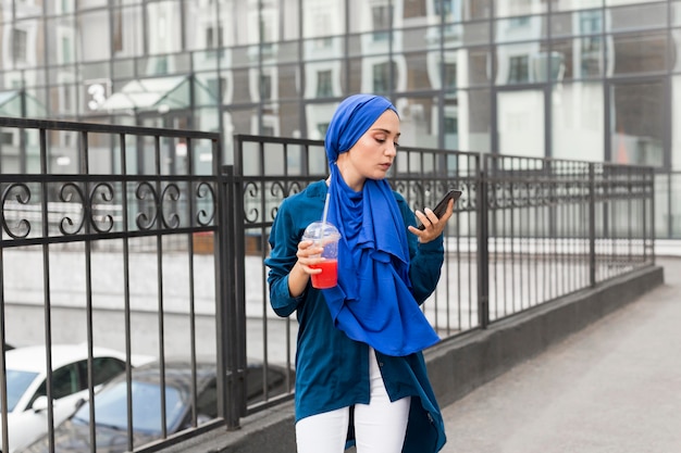Mädchen, das einen Hijab trägt und einen Smoothie hält, während sie ihr Telefon betrachtet