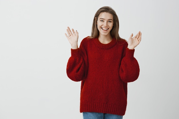 Mädchen, das Freunde über Verlobung erzählt. Porträt der aufgeregten erfreuten bezaubernden Frau im schönen roten losen Pullover, die Handflächen mit Ring am Finger erhebt, jubelt und Glück über graue Wand ausdrückt