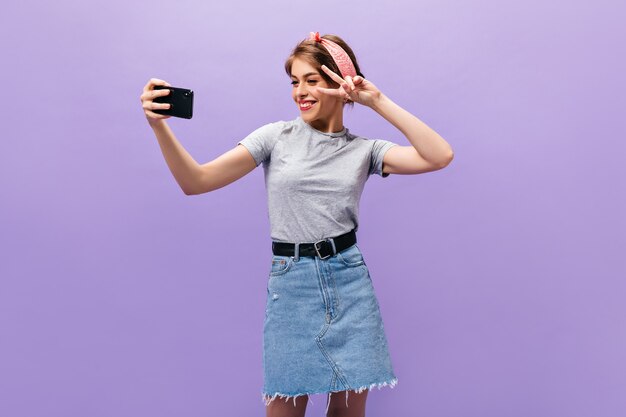 Mädchen, das Friedenszeichen zeigt, zwinkert und Selfie auf lila Hintergrund nimmt. Wunderbare junge Frau im modischen Outfit posiert.