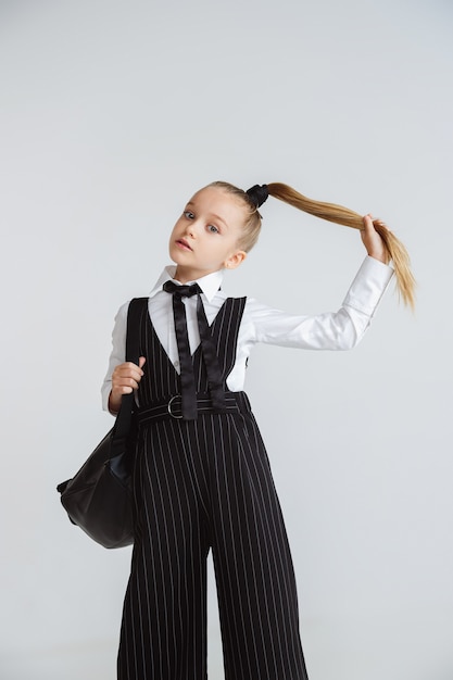 Kostenloses Foto mädchen, das für schule nach einer langen sommerpause vorbereitet. zurück zur schule. kleines weibliches kaukasisches modell, das in der schuluniform mit rucksack auf weißer wand aufwirft. konzept für kindheit, bildung, urlaub.