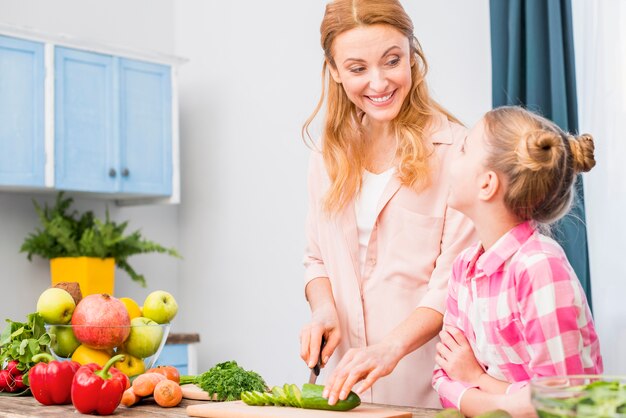 Mädchen, das ihre lächelnde Mutter schneidet die Gurke mit Messer in der Küche betrachtet