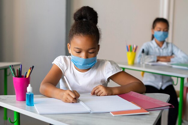 Mädchen, das in der Klasse schreibt, während es eine medizinische Maske trägt