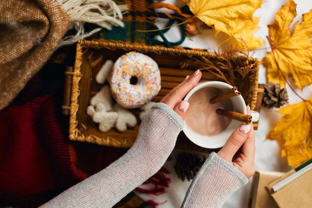 Mädchen, das leckeres Frühstück im Bett auf Holztablett mit Tasse Kakao, Zimt, Keksen und glasierten Donuts isst.