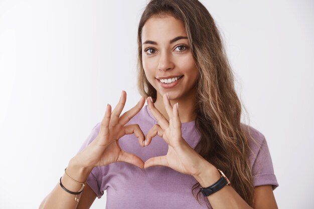 Mädchen, das schlüssel ihr herz sucht. attraktive weibliche, zart gebräunte europäische frau lockiges haar, die liebesgeste zeigt, die breit lächelt und sympathie-romantik ausdrückt, beichtet, stehende weiße wand