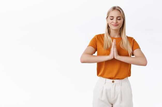 Mädchen, das sich beim Morgenyoga entspannt. Attraktive blonde Frau in orangefarbenem T-Shirt drückt die Handflächen über der Brust zusammen, um zu meditieren, lächelt erfreut, schließen die Augen, führt Atemübungen durch, weißer Hintergrund