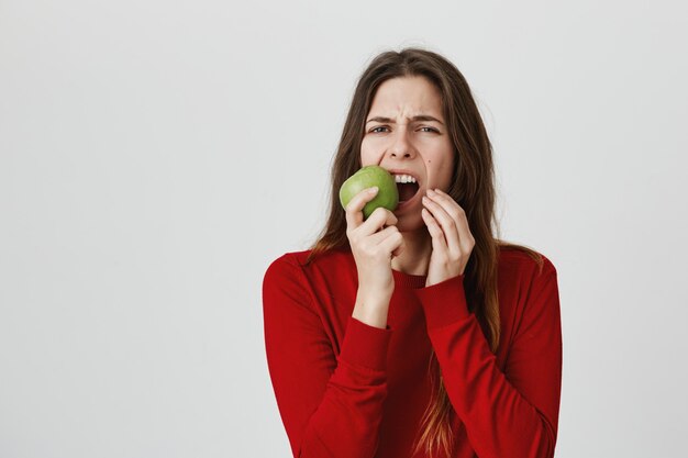 Mädchen, das Zahnschmerzen und Grimassenbildung vom Schmerz als beißenden grünen Apfel fühlt