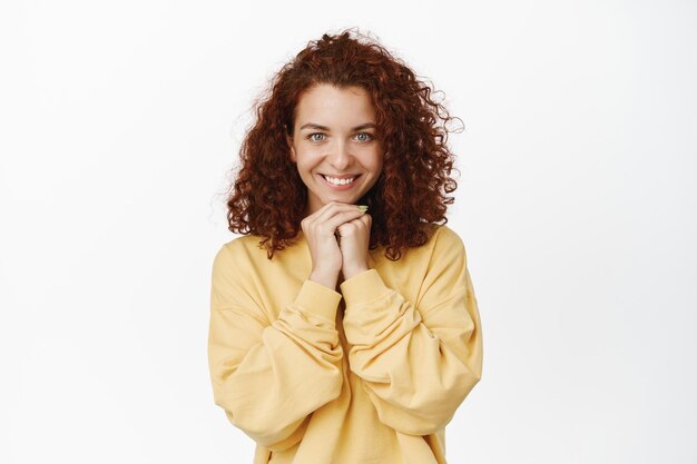 Mädchen erwartet etw. Junge, hoffnungsvolle, lockige Frau, die wartet, lächelt und hoffnungsvoll aussieht, um etwas bettelt und in gelbem Sweatshirt auf weißem Hintergrund steht.