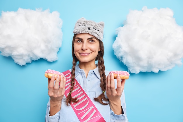 Mädchen feiert Geburtstag hält zwei glasierte Donuts Träume von angenehmen Dingen in Freizeitkleidung isoliert auf Blau on