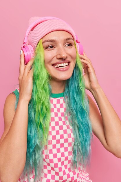 Mädchen genießt Audiospur in drahtlosen Kopfhörern chillt drinnen hört Musik mit lautem Ton hat bunt gefärbte Haare trägt Hut und kariertes Kleid isoliert auf Rosa
