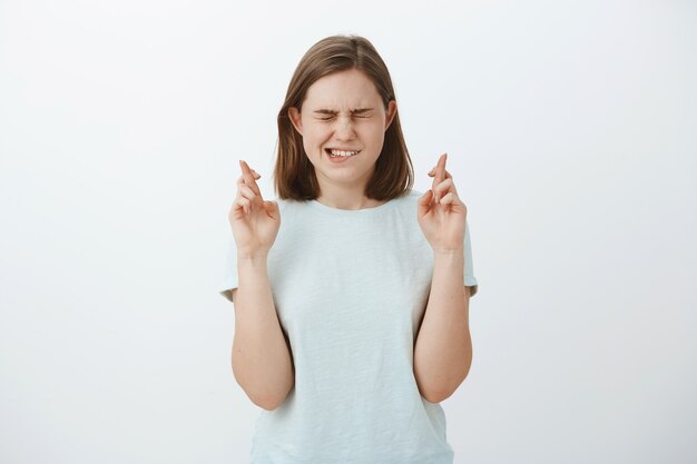 Mädchen glauben, dass Wunder passieren kann, wenn es schwer ist. Intensive hoffnungsvolle charmante Frau mit niedlichen Augen, die das Ohr schließen, und beißenden Unterlippen, die den Wunsch nach Traum haben, werden wahr, wenn sie die Finger drücken, um Glück zu haben