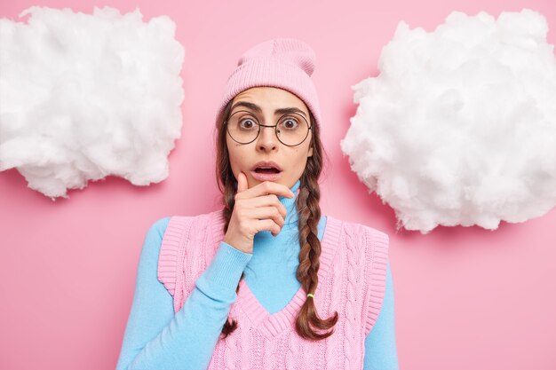 Mädchen hält Kinn steht sprachlos drinnen trägt runde transparente Brille Freizeitkleidung reagiert auf unglaubliche Nachrichtenposen gegen rosig