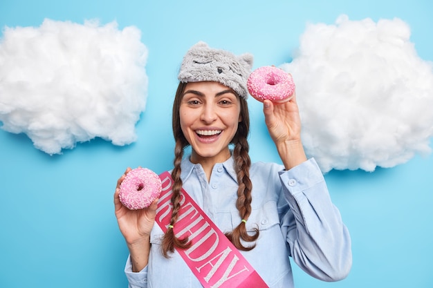 Mädchen hat zwei zöpfe hat fröhliche stimmung trägt schlafmaskenhemd lächelt angenehm posiert mit donuts genießt es, leckeres dessert zu besonderen anlässen einzeln auf blau zu essen