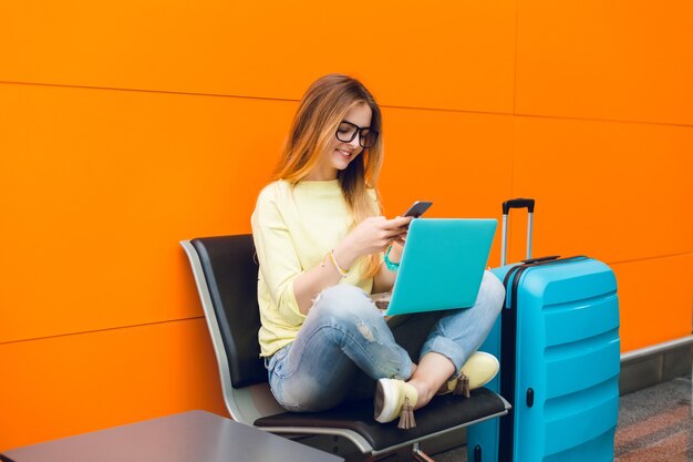 Mädchen im gelben Pullover und in den blauen Jeans sitzt auf Stuhl auf orange Hintergrund. Sie hat einen großen Koffer in der Nähe und einen Laptop auf den Knien. Sie tippt am Telefon.