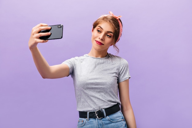 Mädchen im grauen Hemd, das lächelt und selfie auf lila Hintergrund nimmt. Schöne trendige Frau mit rosa Stirnband in coolen Kleidern macht Foto.