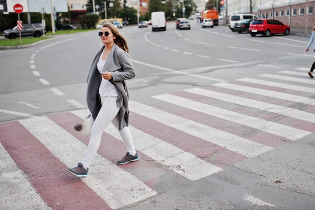 Mädchen im grauen Mantel mit Sonnenbrille und Handtasche, das auf dem Fußgängerüberweg geht