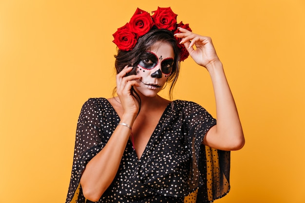 Mädchen im Kleid mit V-Ausschnitt und weiten Ärmeln wirft für Porträt auf isolierter Wand auf. Mexikanisches Mädchen mit Rosen auf dem Kopf