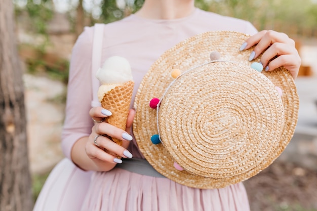 Mädchen im niedlichen lila Kleid, das im Freien steht und dekorierten Strohsommerhut in der Hand hält