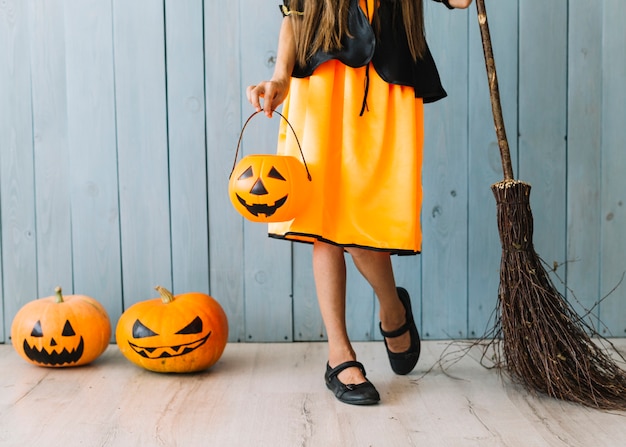 Mädchen im orange und schwarzen Kleid, das mit Halloween-Korb und -besen steht