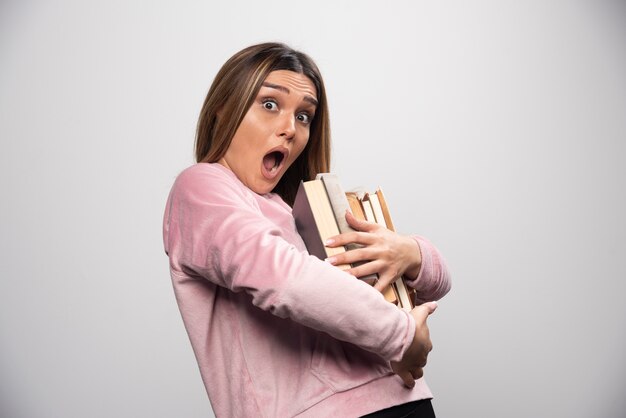 Mädchen im rosa Swaetshirt, das schweren Stapel Bücher hält und trägt.