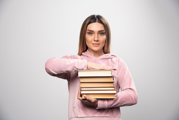 Mädchen im rosa Sweatshirt, das schweren Stapel Bücher hält und trägt.