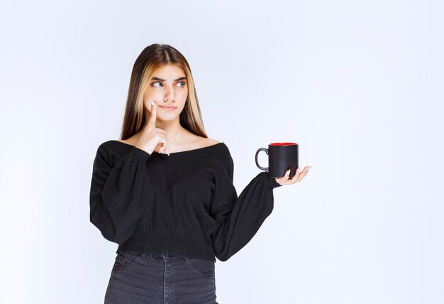 Mädchen im schwarzen Hemd, das eine schwarze Kaffeetasse hält und denkt. Foto in hoher Qualität