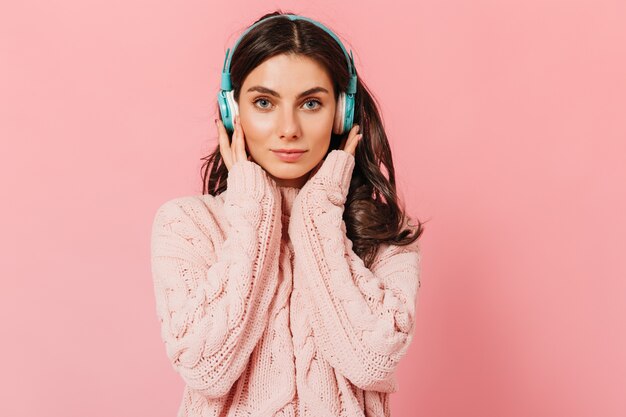Mädchen im Strickpullover drückt Kopfhörer für besseren Klang. Blauäugige Frau mit leichtem Lächeln schaut in Kamera auf rosa Hintergrund.