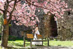 Kostenloses Foto mädchen im weißen kleid steht unter rosa blühendem kirschblüte-baum im park