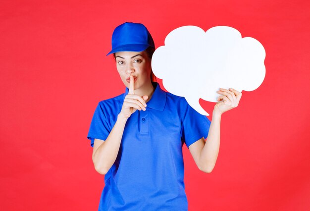 Mädchen in blauer Uniform und Baskenmütze, das ein Denkbrett in Wolkenform hält und um Stille bittet.