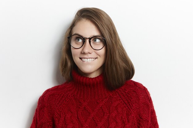 Mädchen in Brille und gemütlichem warmen Pullover schaut auf und beißt sich auf die Lippen, als hätte es eine tolle Idee. Menschen, Stil, Optik, Vision, Brille und Modekonzept