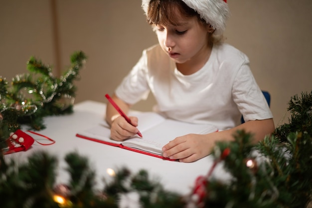 Mädchen in einem weißen t-shirt mit einem roten stift und einer neujahrsmütze auf dem kopf schreibt dem lieben weihnachtsmann einen brief an den tisch in erwartung der geburt. wunschliste mit geschenken für das neue jahr.