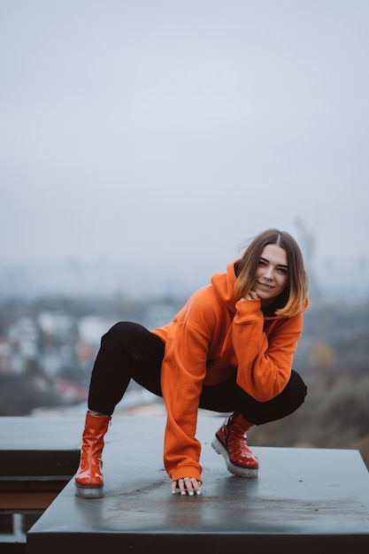 Mädchen in einer orange Jacke posiert auf dem Dach eines Gebäudes im Stadtzentrum