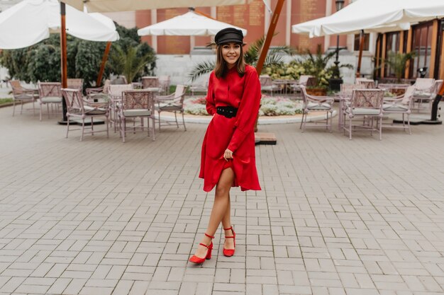 Mädchen in elegantem, rotem Gürtelkleid und Schuhen auf Stadtabsatz zeigt schlanke Beine. Foto in voller Länge im Stadtcafé