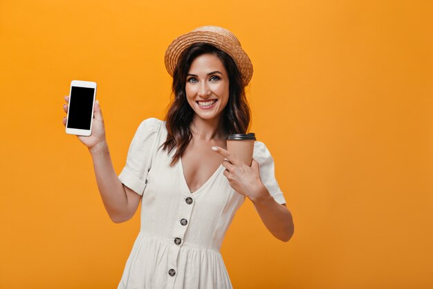 Mädchen in Hut und Kleid zeigt auf Smartphone und hält Glas Tee