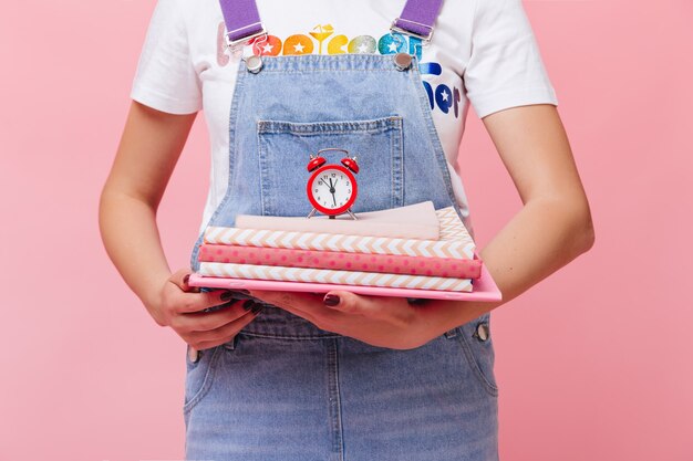 Mädchen in Jeans-Overalls hält rosa Notizbücher und Wecker