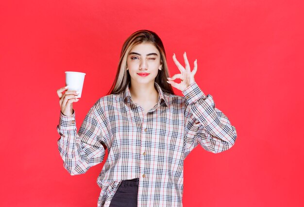 Mädchen in kariertem Hemd, das eine weiße Einweg-Kaffeetasse hält und den Geschmack genießt