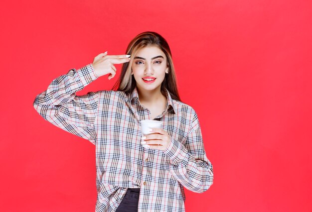 Mädchen in kariertem Hemd, das eine weiße Einweg-Kaffeetasse hält und nachdenklich aussieht oder gute Ideen hat