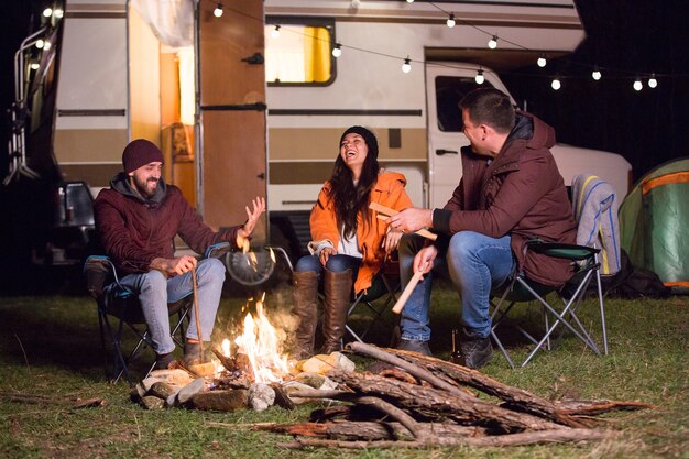 Mädchen lacht hart, nachdem ihre Freunde einen Witz am Lagerfeuer mit Retro-Wohnmobil im Hintergrund erzählt haben.