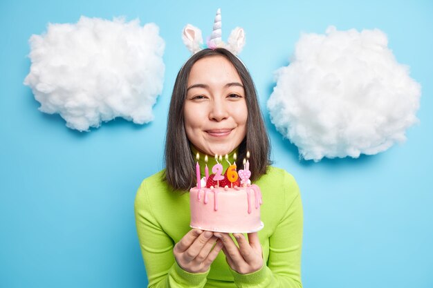 Mädchen lächelt sanft hält köstlichen Kuchen mit Kerzen lädt Sie zur Party ein, schaut glücklich in die Kamera posiert drinnen auf Blau