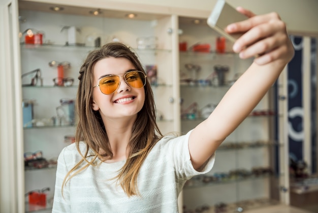 Mädchen lebt nie zu Hause ohne Smartphone, stilvolle europäische Brünette, die Sonnenbrille anprobiert, während Selfie am Telefon nimmt, breit lächelnd