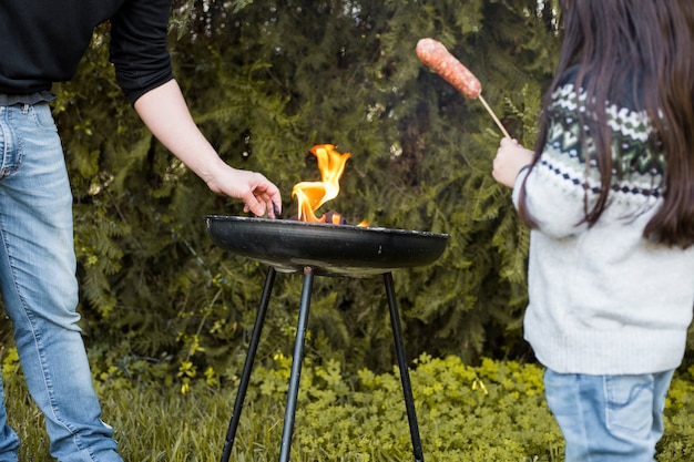 Mädchen mit der Wurst, die nahe ihrem Vater grillen auf tragbarem Grill steht