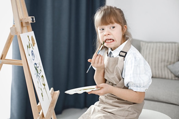 Mädchen mit Down-Syndrom, das eine beige Schürze trägt und auf einer Staffelei malt