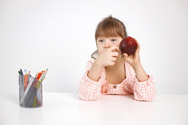 Mädchen mit Down-Syndrom sitzt am Schreibtisch und hält einen Apfel