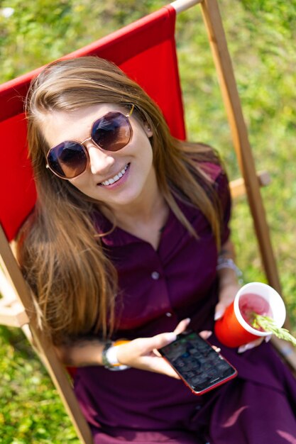 Mädchen mit einem Smartphone und einem Cocktail in der Natur in einem Liegestuhl. Ruhetag im Sommer.