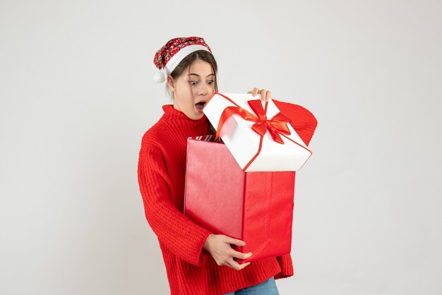 Mädchen mit großen Augen und Weihnachtsmütze, das ihr Weihnachtsgeschenk auf Weiß öffnet