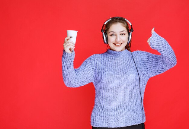 Mädchen mit Kopfhörern, die eine Kaffeetasse halten und Genusszeichen zeigen.