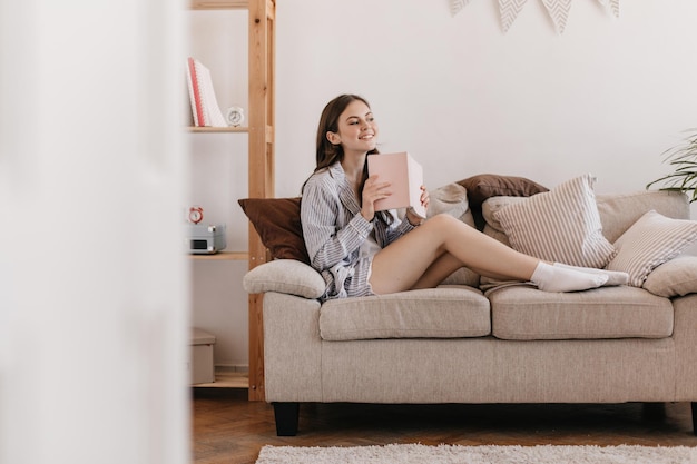 Mädchen mit Lächeln posiert auf der Couch sitzend Frau im Schlafanzug mit Notizblock Verträumter Teenager in gestreiftem blauem Hemd hält rosa Buch und hat coole Zeit zu Hause