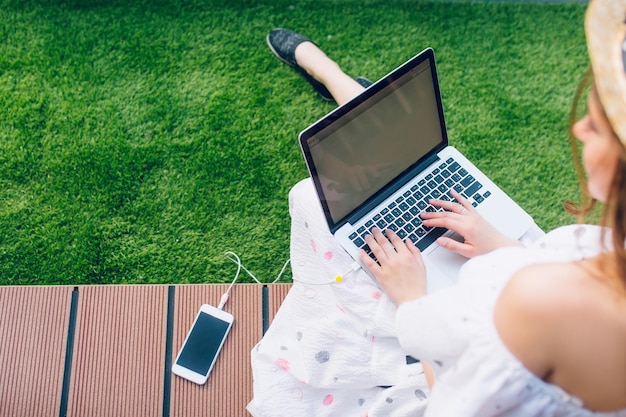 Mädchen mit langen Haaren im Hut sitzt auf dem Dielenboden. Sie trägt ein weißes Kleid mit nackten Schultern. Sie tippt auf Knien auf einem Laptop. Ansicht von oben, Fokus auf Laptop